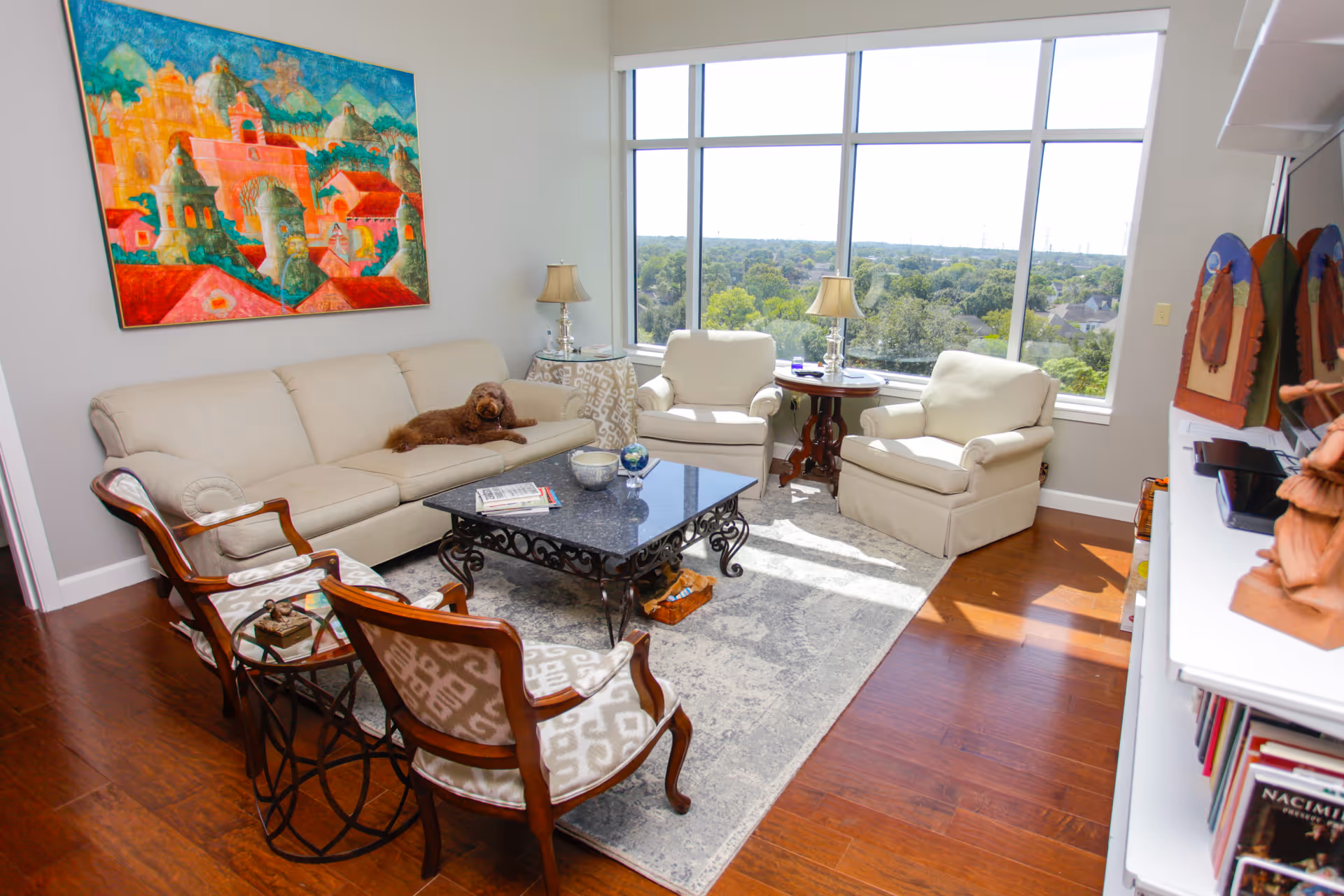 A bright living room with large windows overlooking greenery. The room features a beige sofa with a small brown dog lying on it, two beige armchairs, two wooden chairs with patterned cushions, a black ornate coffee table, a side table with a lamp, and a colorful painting on the wall. Hardwood floors and a light-colored area rug complete the space.