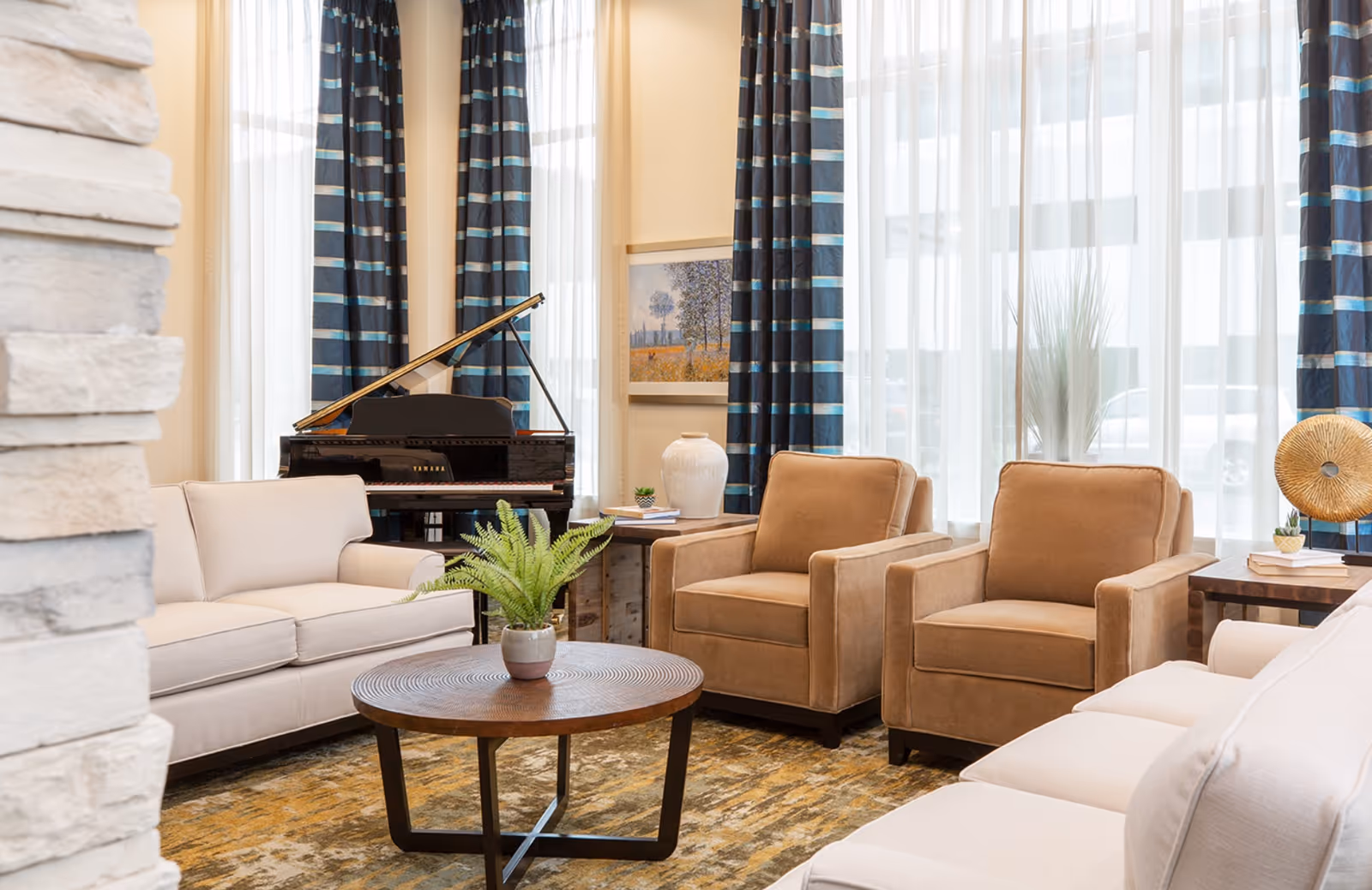 A bright and cozy living room area with beige sofas and brown armchairs arranged around a round wooden coffee table with a potted plant. In the background, there is a black Yamaha grand piano, large windows with sheer white curtains and blue striped drapes, a framed artwork on the wall, and decorative items on side tables.