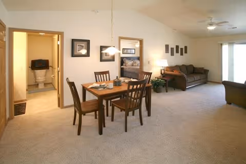 Interior view of a senior living facility apartment showing a dining area with a wooden table and four chairs, a living room with a sofa and armchair, and a bedroom visible through an open doorway. A bathroom is also visible through another open door. The space is carpeted and well-lit with ceiling lights and a lamp.