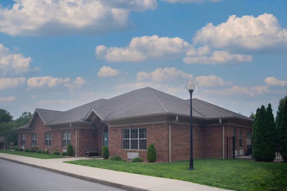 Exterior view of a single-story brick building with multiple windows, a gray shingled roof, a sidewalk, a street lamp, and neatly trimmed bushes and grass under a partly cloudy sky.