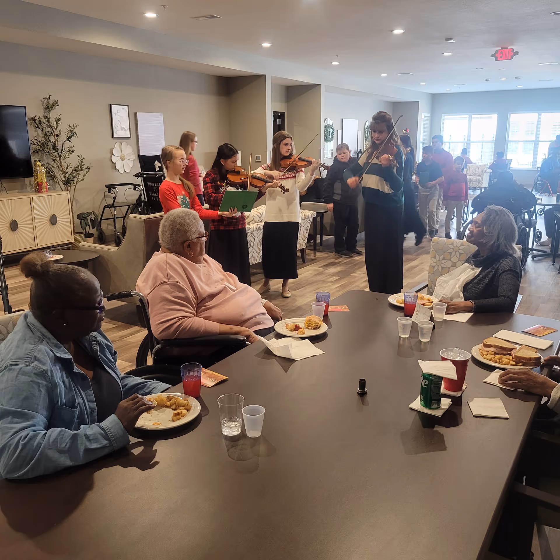 Residents sit around a dining table eating while a small group of young musicians play violins in a common area of the assisted living facility.