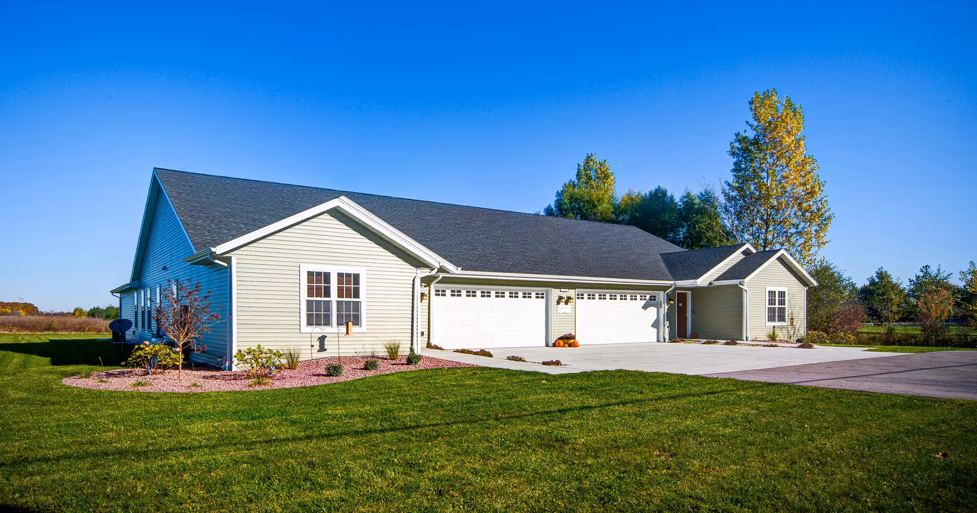 Single-story residential building with light-colored siding, a dark shingled roof, and a three-car garage. The building is surrounded by a well-maintained lawn and some small trees and shrubs. The sky is clear and blue.