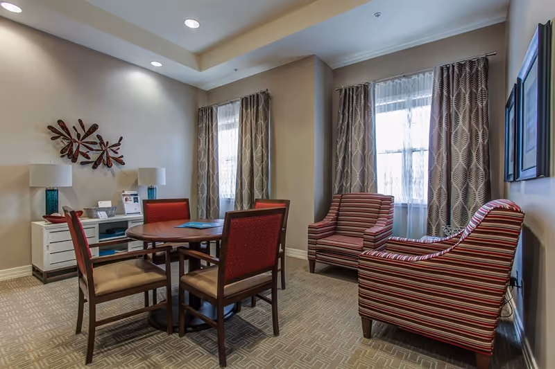 A cozy sitting room with a round table and four chairs, two striped armchairs, a sideboard with lamps, and patterned curtains over two windows.
