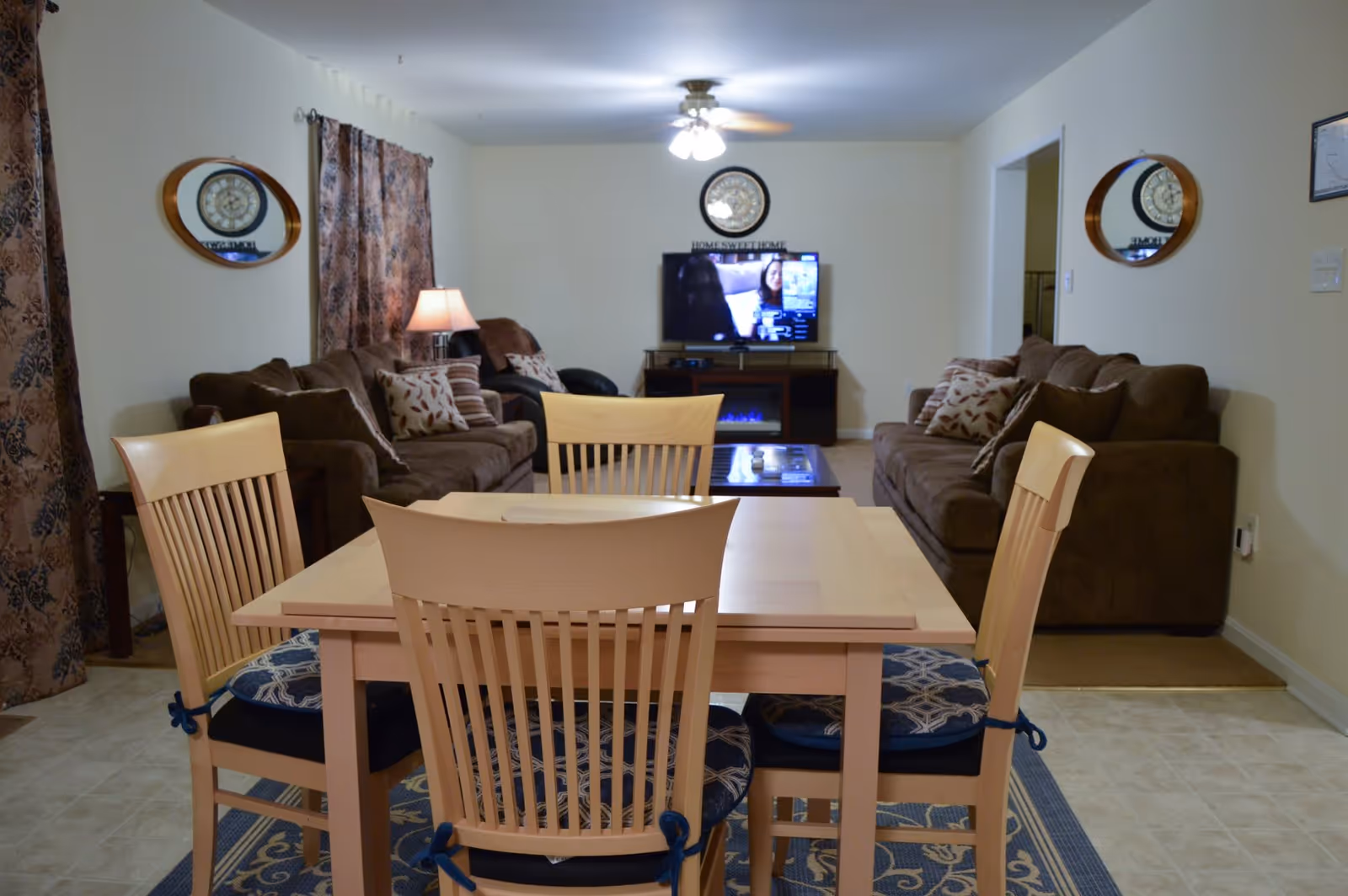 Open-plan living and dining area with a light wood dining table and chairs in front of brown sofas, a TV, and decorative mirrors on the walls.