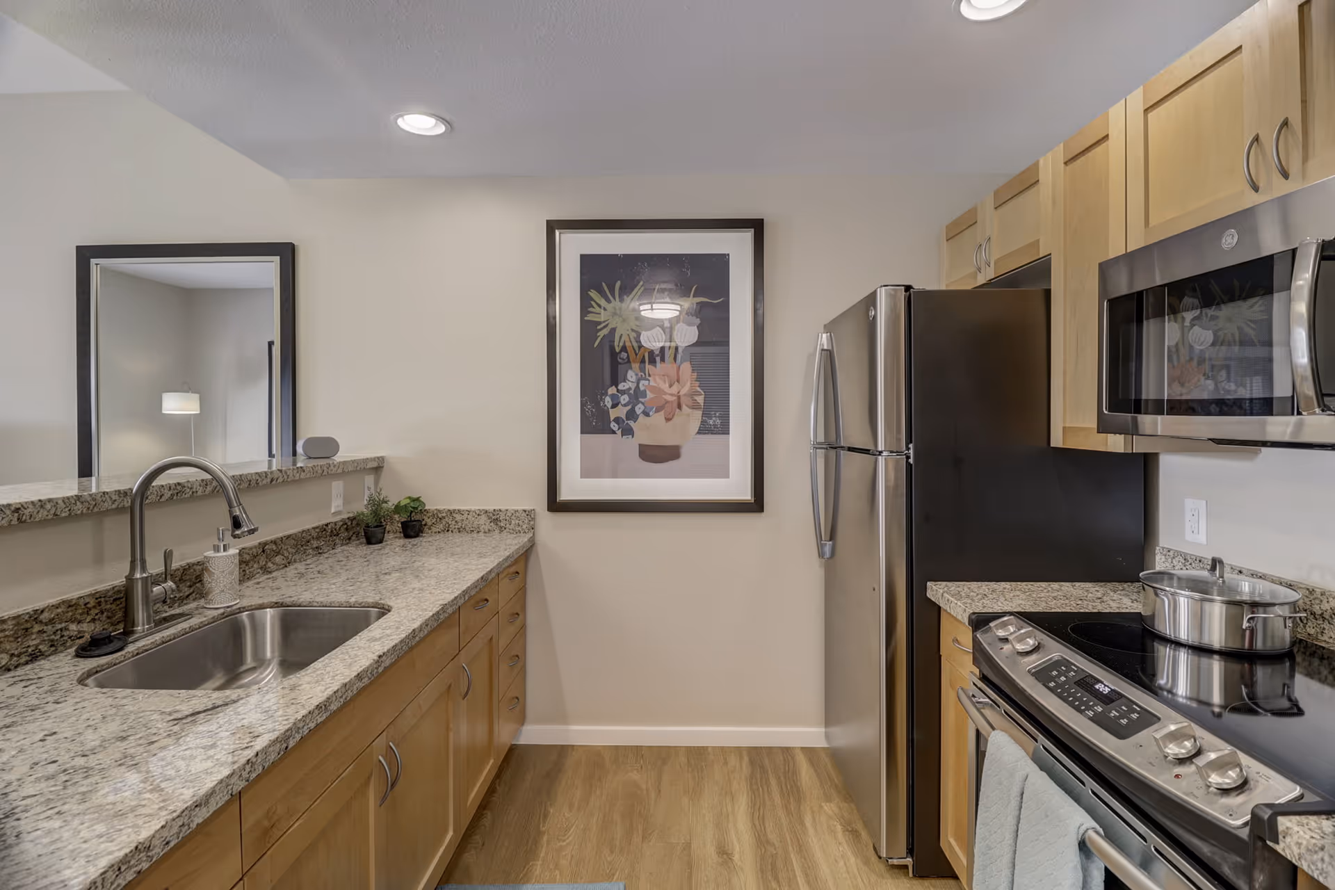 A modern kitchen with light wood cabinets, granite countertops, a stainless steel refrigerator, microwave, and stove. There is a sink with a faucet on the left countertop, two small potted plants, and a framed floral artwork on the wall at the end of the kitchen. The floor has a wood-like finish.