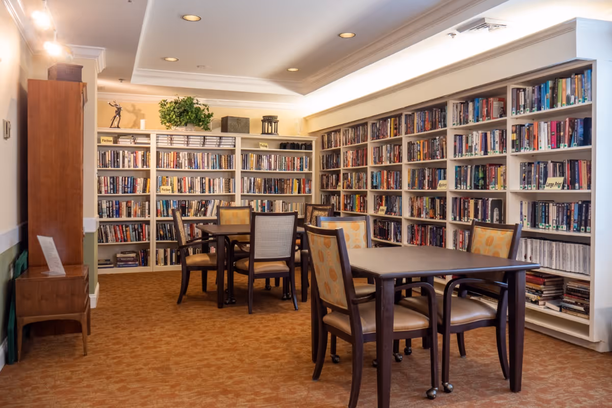 A cozy library room with multiple bookshelves filled with books along the walls. There are two tables with chairs around them for reading or studying. The room has warm lighting and a carpeted floor, creating a comfortable and inviting atmosphere.