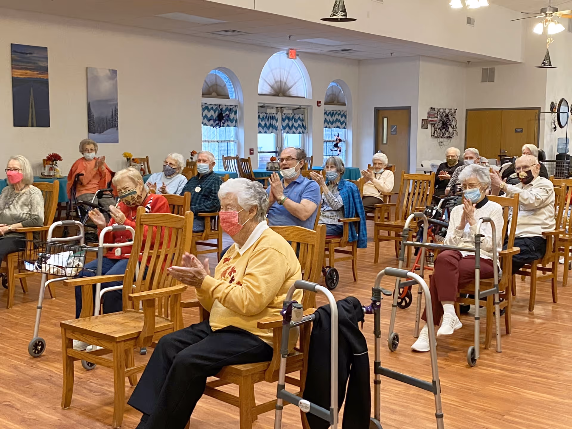 A group of elderly people sitting on wooden chairs in a spacious room with wooden floors, clapping and wearing face masks. Some have walkers beside them. The room has arched windows with patterned curtains and ceiling fans.