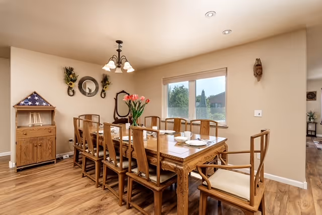 A dining room with a long wooden table surrounded by ten wooden chairs with cushions. The table is set with white plates and bowls, and a vase with pink flowers is placed in the center. The room has light-colored walls, a window with a view outside, a chandelier with five lights hanging above the table, and decorative items on the walls including a round mirror and floral arrangements. The floor is wooden.