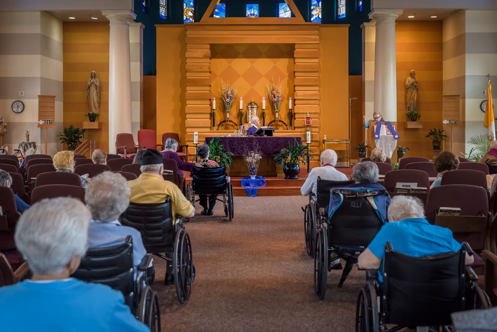 A group of elderly people, many in wheelchairs, seated in rows facing an altar in a chapel. A priest is standing on the right side near the altar, conducting a service. The chapel has stained glass windows, statues, and plants decorating the space.
