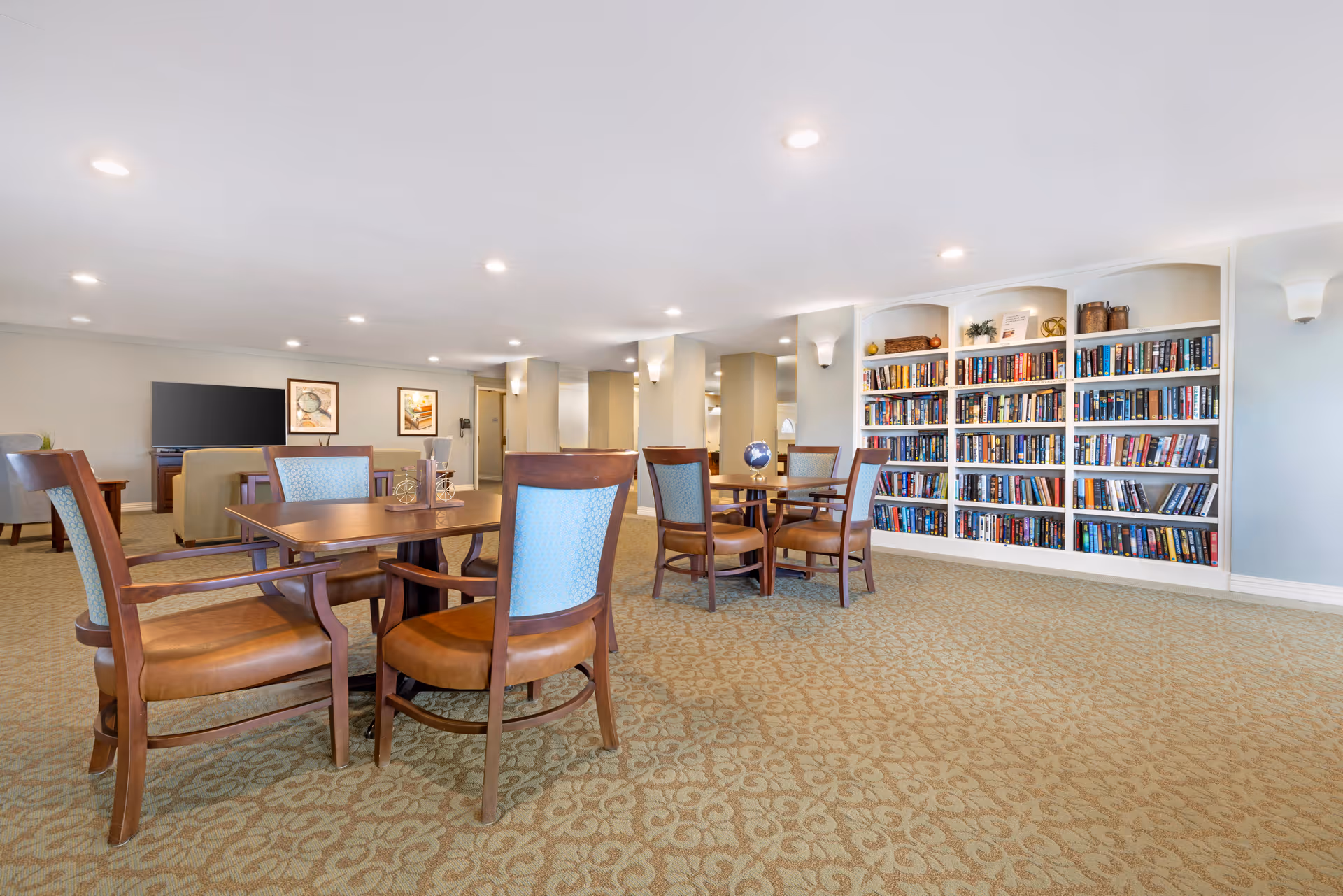 A spacious common area in a senior living facility featuring multiple wooden tables with cushioned chairs arranged around them. The room has a patterned carpet, built-in bookshelves filled with books, and a seating area with a television and framed artwork on the walls. The space is well-lit with recessed ceiling lights and wall sconces.