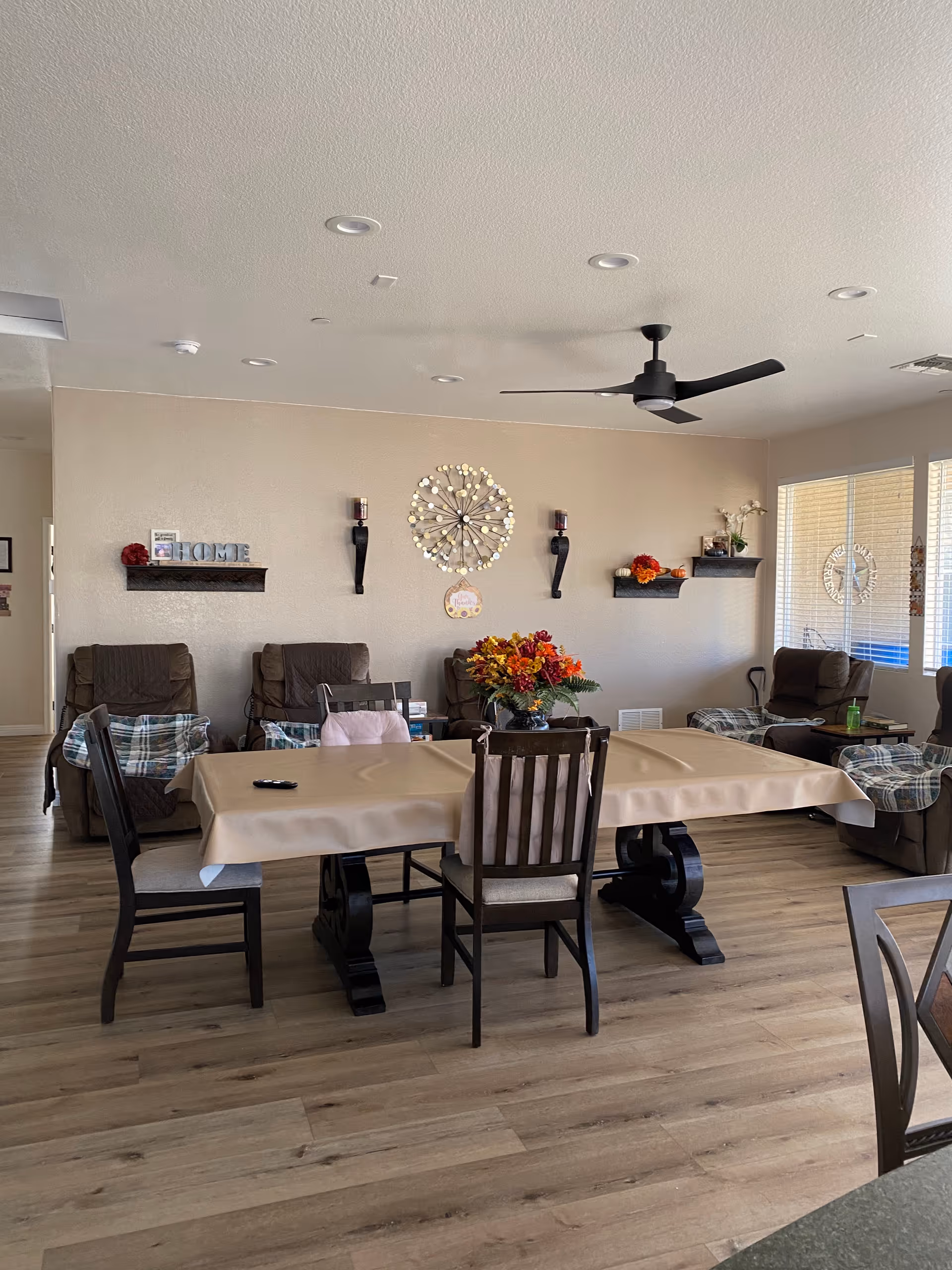 A cozy common area in Silver Creek Assisted Living featuring a rectangular dining table covered with a beige tablecloth and surrounded by wooden chairs. Behind the table are several cushioned recliners with plaid blankets draped over them. The beige wall has decorative shelves with small items and a large circular wall decoration. A ceiling fan with black blades is mounted on the white ceiling. Large windows on the right side let in natural light.