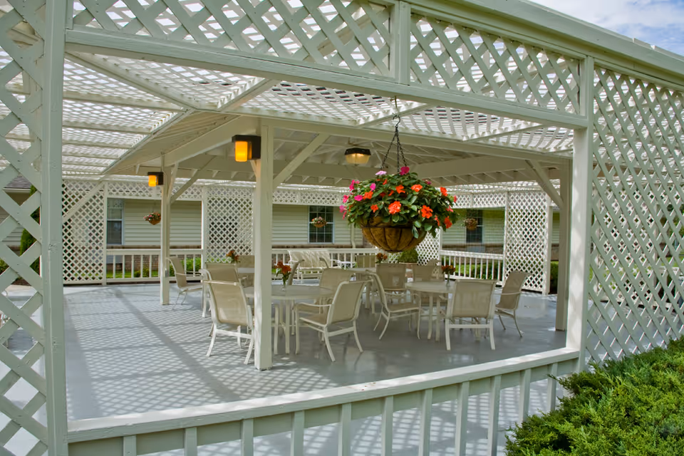 Outdoor covered patio area with white lattice walls and ceiling, furnished with multiple tables and chairs. A hanging basket with vibrant pink and orange flowers is suspended from the ceiling. The patio is part of a residential building with windows visible in the background.