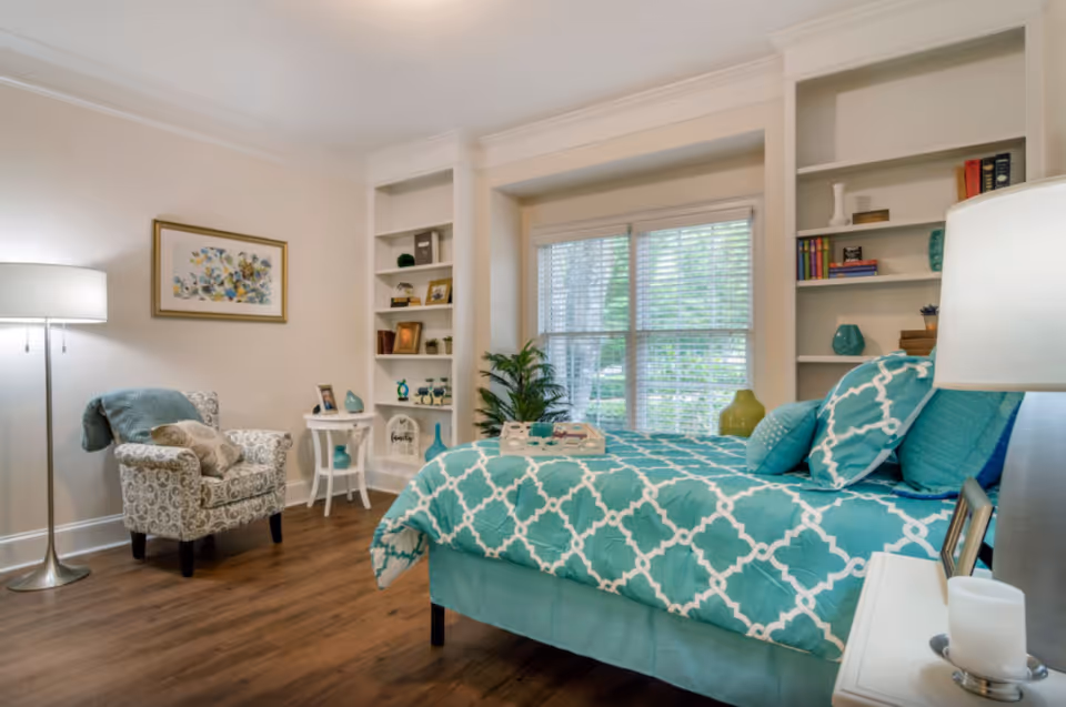 A cozy bedroom with a teal and white patterned bedspread on a bed, a white nightstand with a lamp and candle, a patterned armchair with a teal throw blanket, a small white side table, built-in shelves with decorative items, and a large window with blinds letting in natural light.
