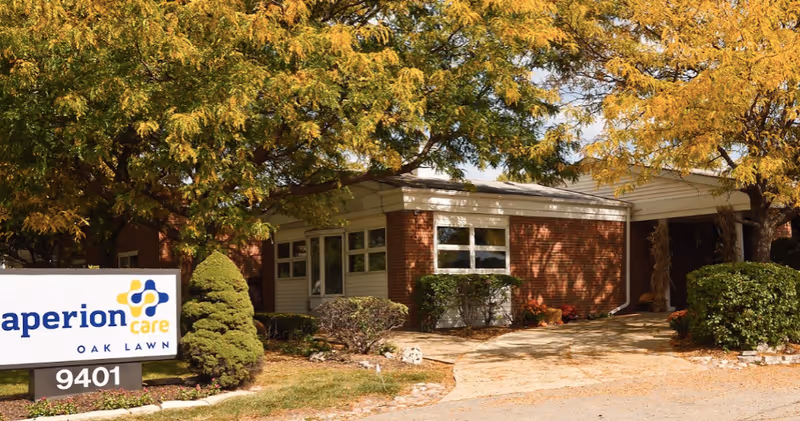 Exterior view of Aperion Care Oak Lawn facility showing a brick building partially obscured by trees with yellow and green leaves. There is a sign in front with the facility name and address number 9401.