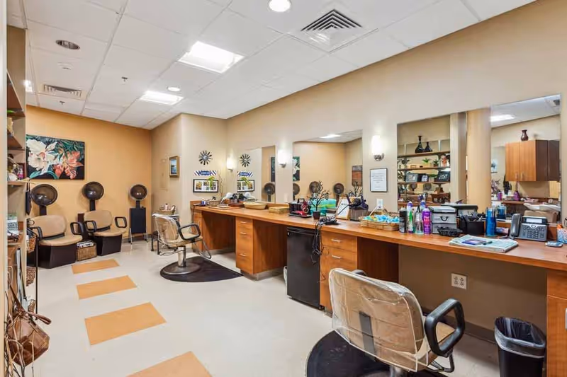 Interior view of a senior living facility salon area with multiple styling stations featuring large mirrors, chairs, and hair care products. There are also three hair drying chairs against the back wall and decorative artwork on the walls.
