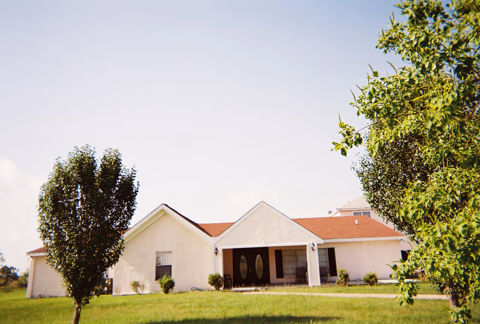 Single-story building with a light-colored exterior and a red roof, surrounded by green grass and trees under a clear sky.