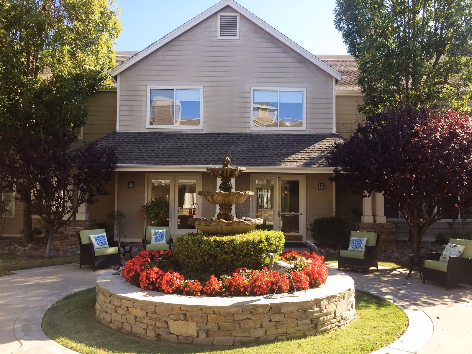 Outdoor courtyard area of a senior living facility with a central stone fountain surrounded by red flowers and green shrubbery. There are four cushioned chairs with blue and white pillows arranged around the fountain, shaded by trees. The building in the background has beige siding, two windows on the upper floor, and glass doors on the ground floor.