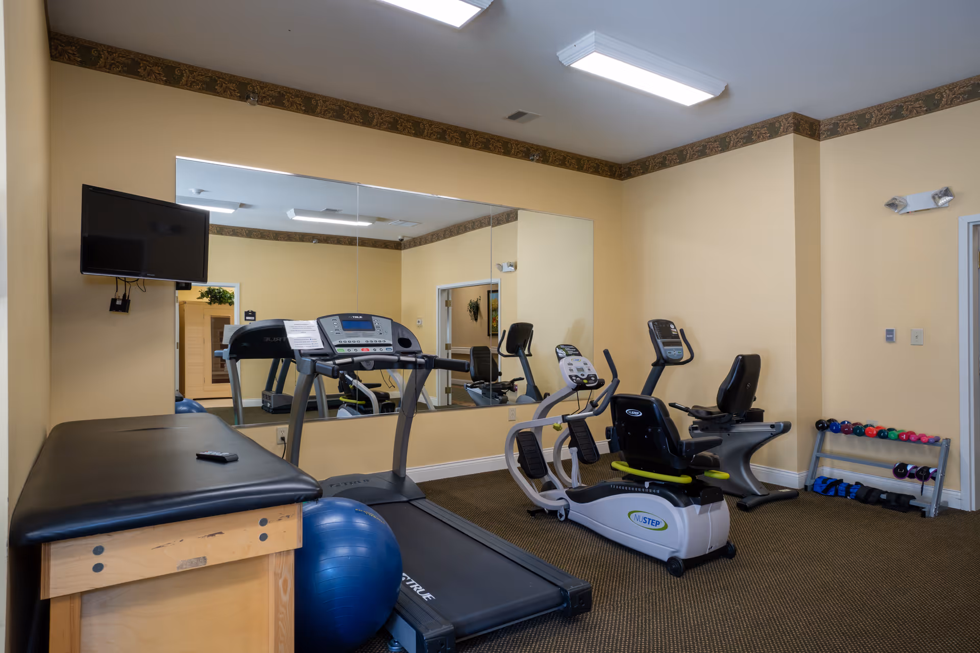 Interior view of a small fitness room with exercise equipment including a treadmill, two recumbent stationary bikes, a blue exercise ball, a padded bench, and a rack of colorful dumbbells. A large mirror covers one wall, reflecting the equipment and room. A wall-mounted TV is visible on the left side.