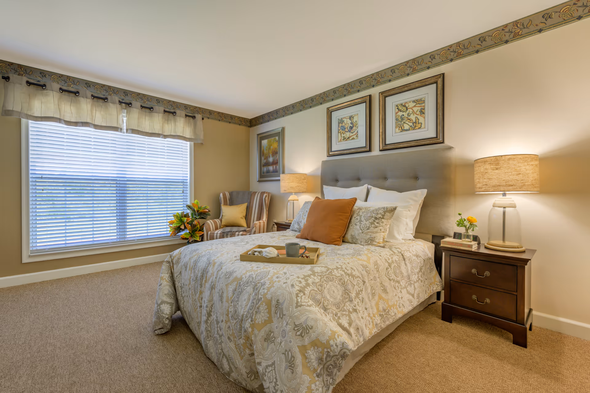 A cozy bedroom with a large window covered by blinds and a valance. The room features a bed with a patterned comforter, multiple pillows, and a tray with a cup and napkin. On either side of the bed are wooden nightstands with lamps and small decorative items. A striped armchair with a yellow pillow and a potted plant are positioned near the window. The walls are decorated with framed artwork and a floral border near the ceiling.
