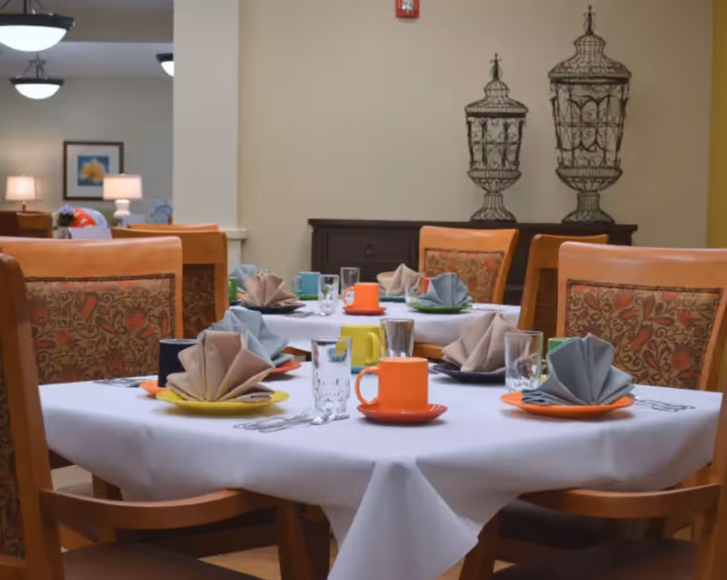 Round dining tables set with colorful plates, folded napkins, glasses and mugs in a senior living dining area.