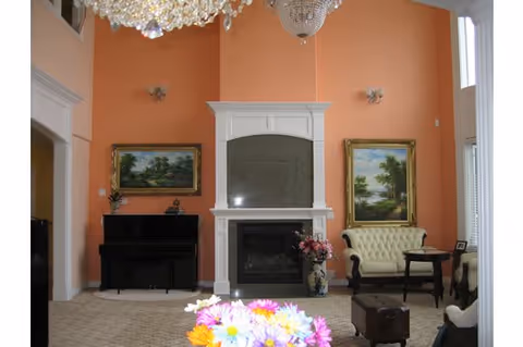 Elegant living room with a chandelier, fireplace flanked by framed landscape paintings, a piano, tufted seating, and a floral centerpiece on a table.