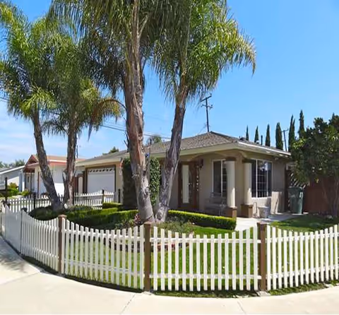 A single-story home-style senior living facility with a white picket fence and palm trees in sunshine.
