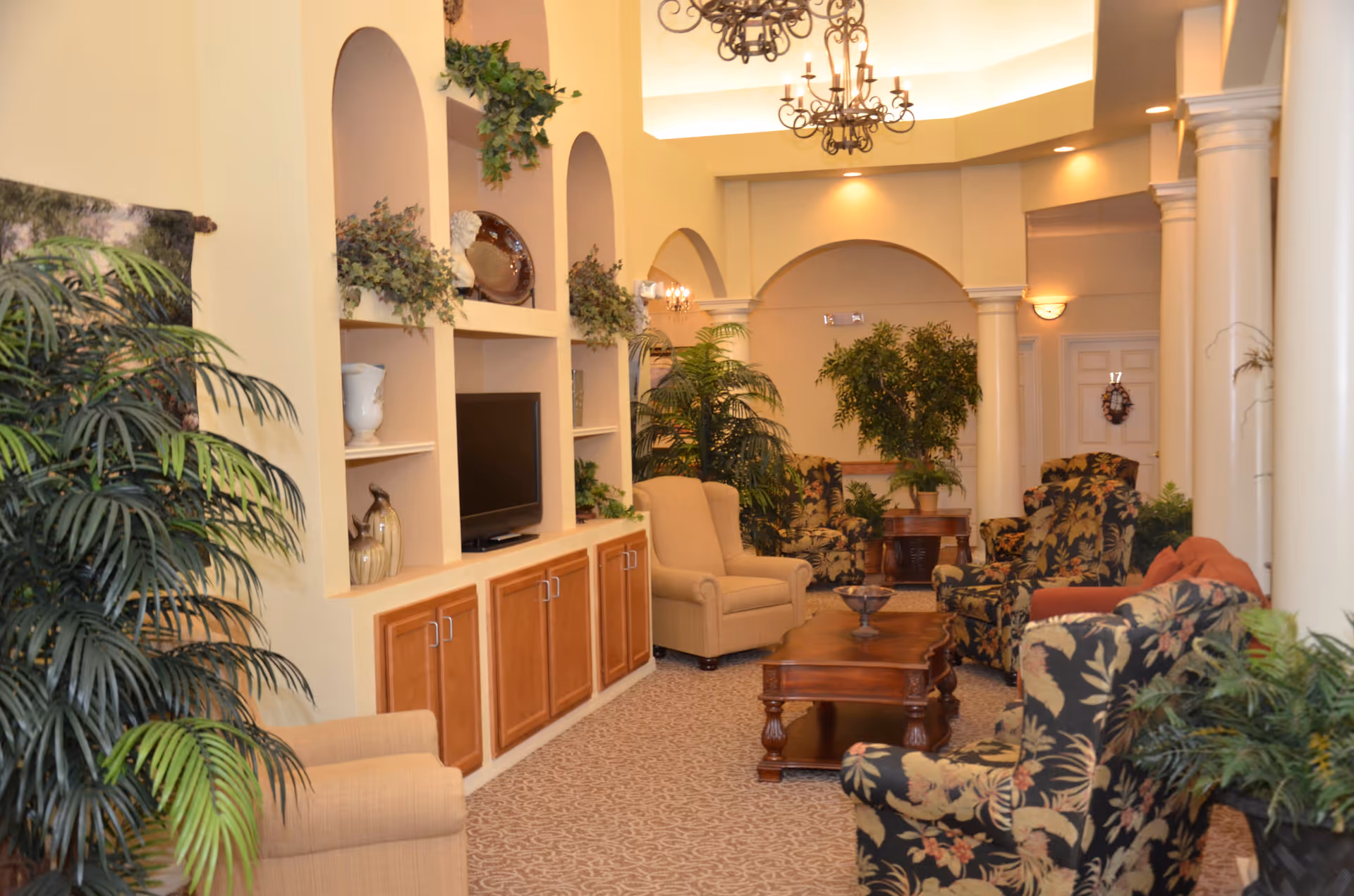 A cozy living room area in a senior living facility with beige walls and carpet. The room features multiple upholstered armchairs with floral patterns, a wooden coffee table, built-in shelves with decorative items, a flat-screen TV, and several potted plants. The ceiling has a chandelier and recessed lighting, with architectural columns and arches adding to the decor.
