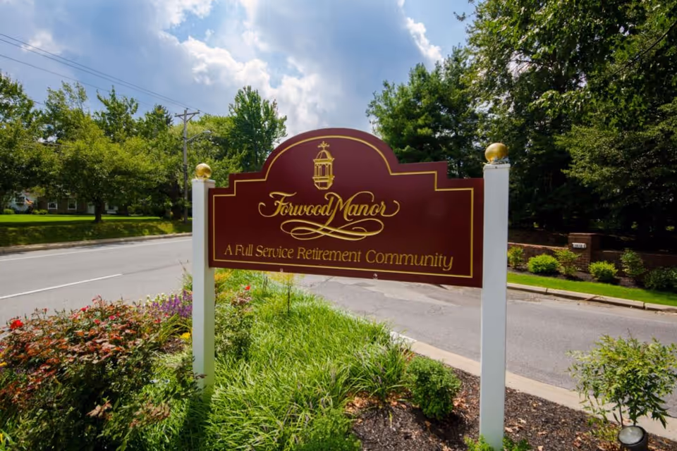 A maroon and gold sign for Forwood Manor, a full service retirement community, standing on a landscaped area with flowers and greenery beside a road with trees and a partly cloudy sky in the background.