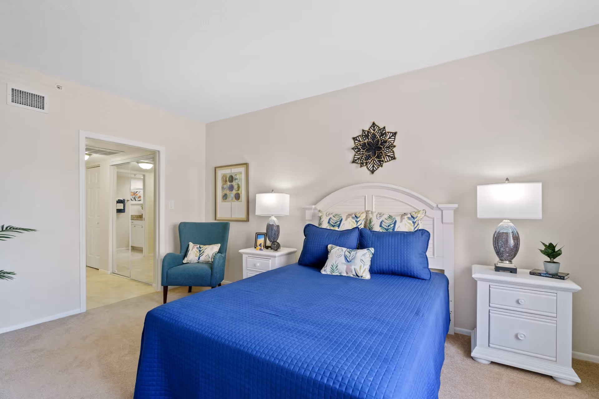 A bedroom with a bed covered in a blue quilt and decorative pillows with leaf patterns. There are two white nightstands on either side of the bed, each with a lamp. A blue armchair with a matching pillow is placed near the doorway, which leads to a bathroom with mirrored closet doors. The walls are light-colored, and there is a decorative wall hanging above the bed.