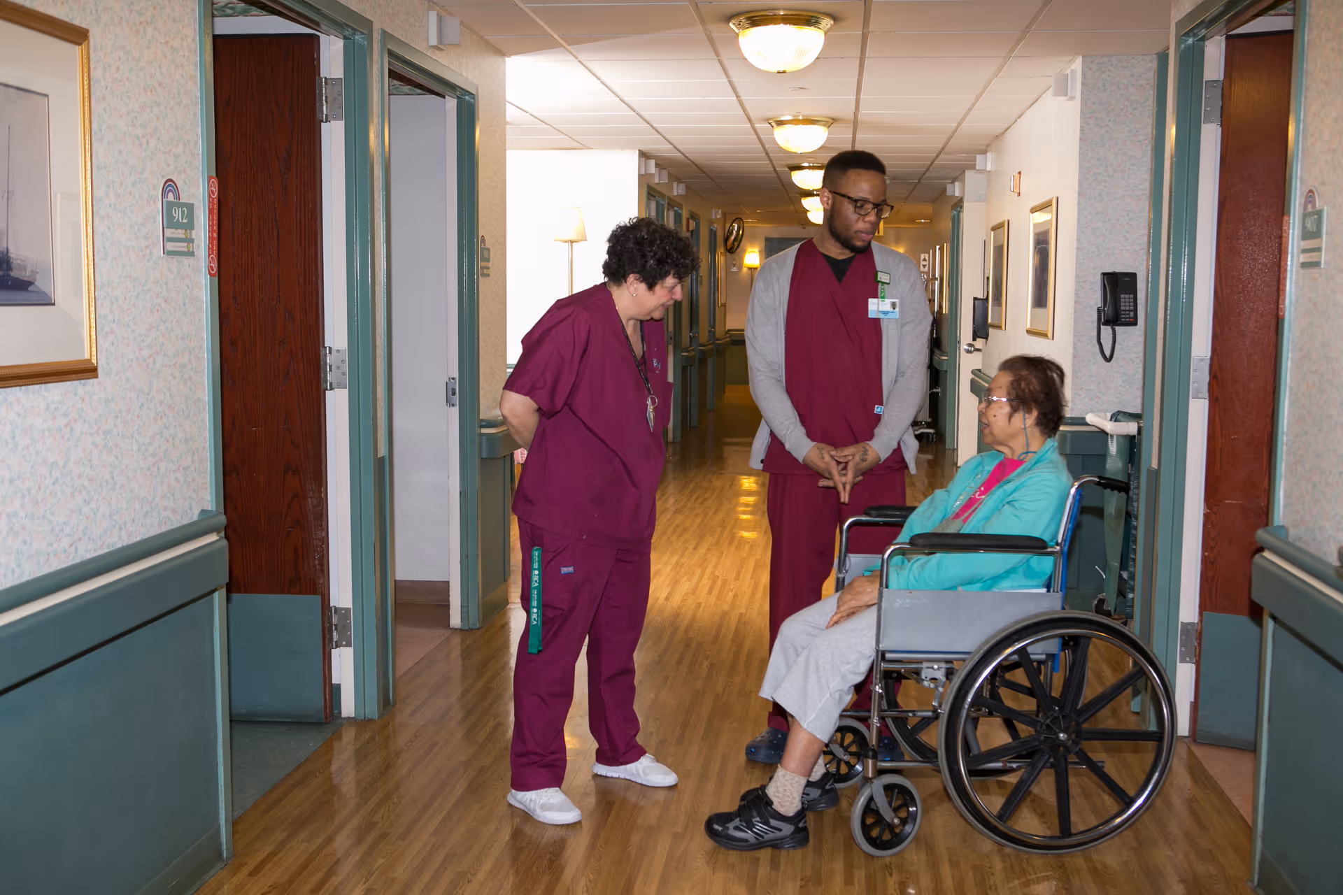 Two healthcare workers in maroon scrubs are standing in a hallway talking to an elderly woman seated in a wheelchair. The hallway has wooden floors, green and beige walls, and several doors and framed pictures along the walls.
