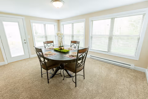 Bright dining area with a round table set for four surrounded by large windows and carpeted floor.