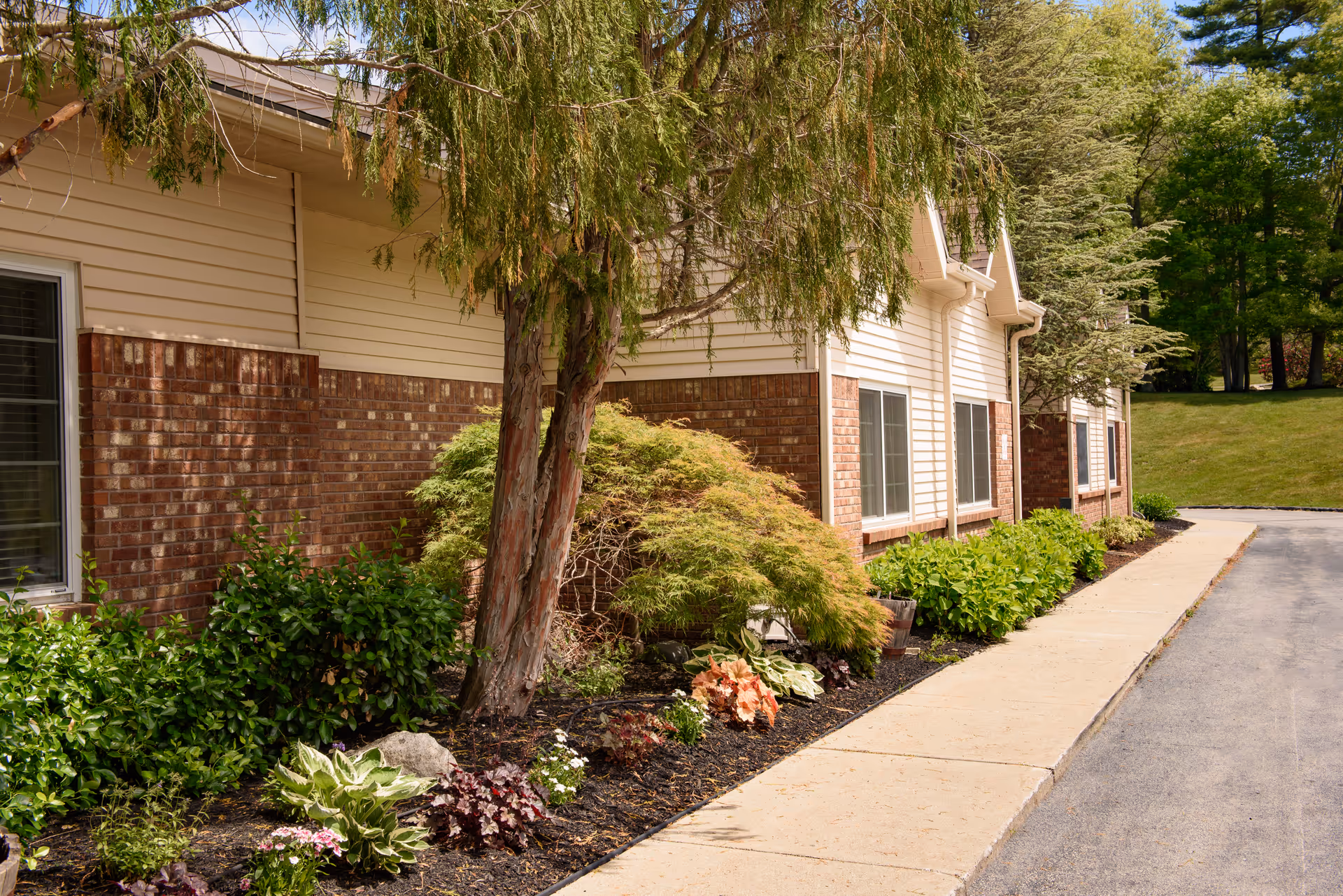 Exterior view of a building with beige siding and red brick lower walls, surrounded by landscaped greenery including bushes, small trees, and flowers along a sidewalk next to a paved driveway.