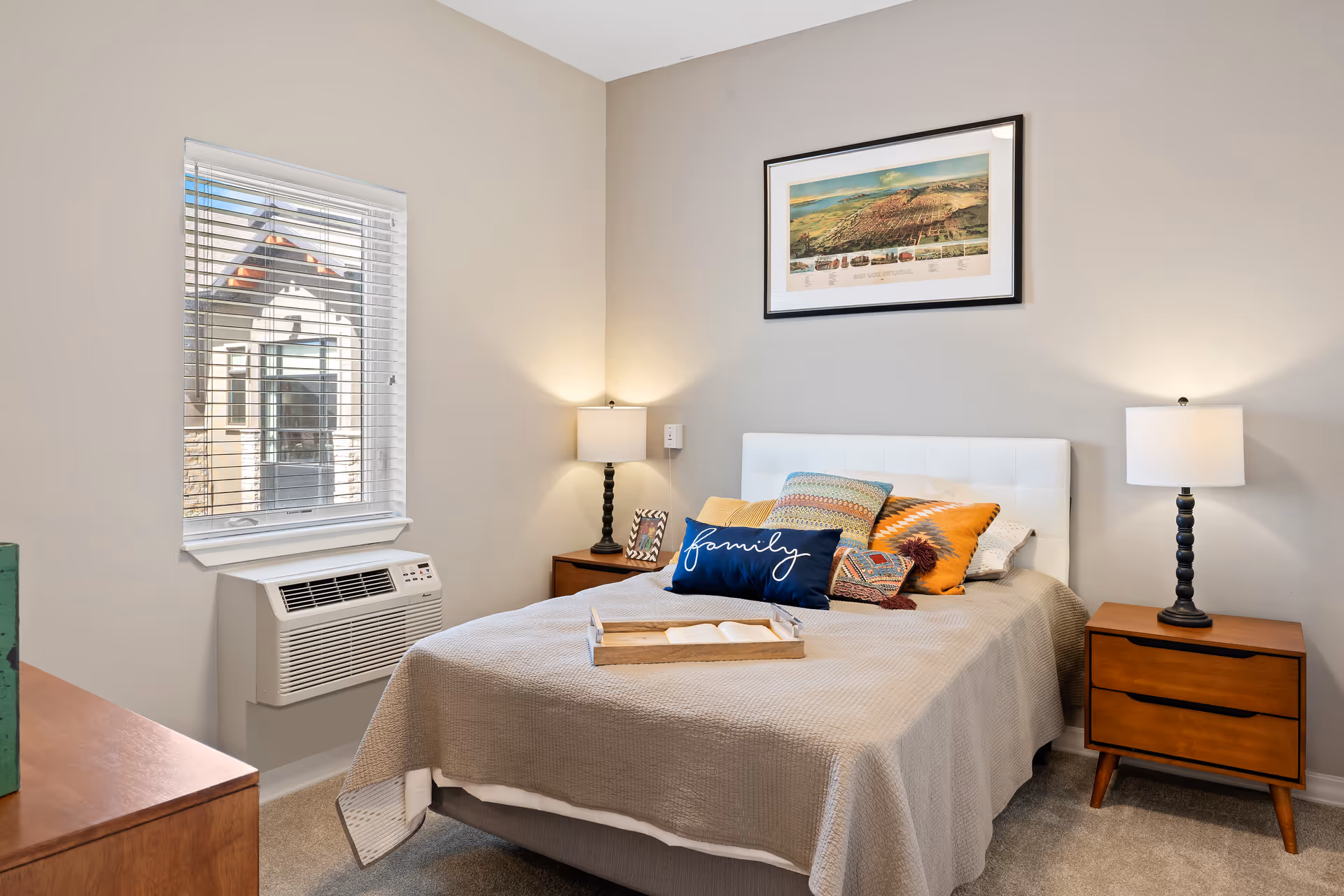 Neatly decorated bedroom with a made bed, colorful pillows, two nightstands with lamps, a window and framed wall art.