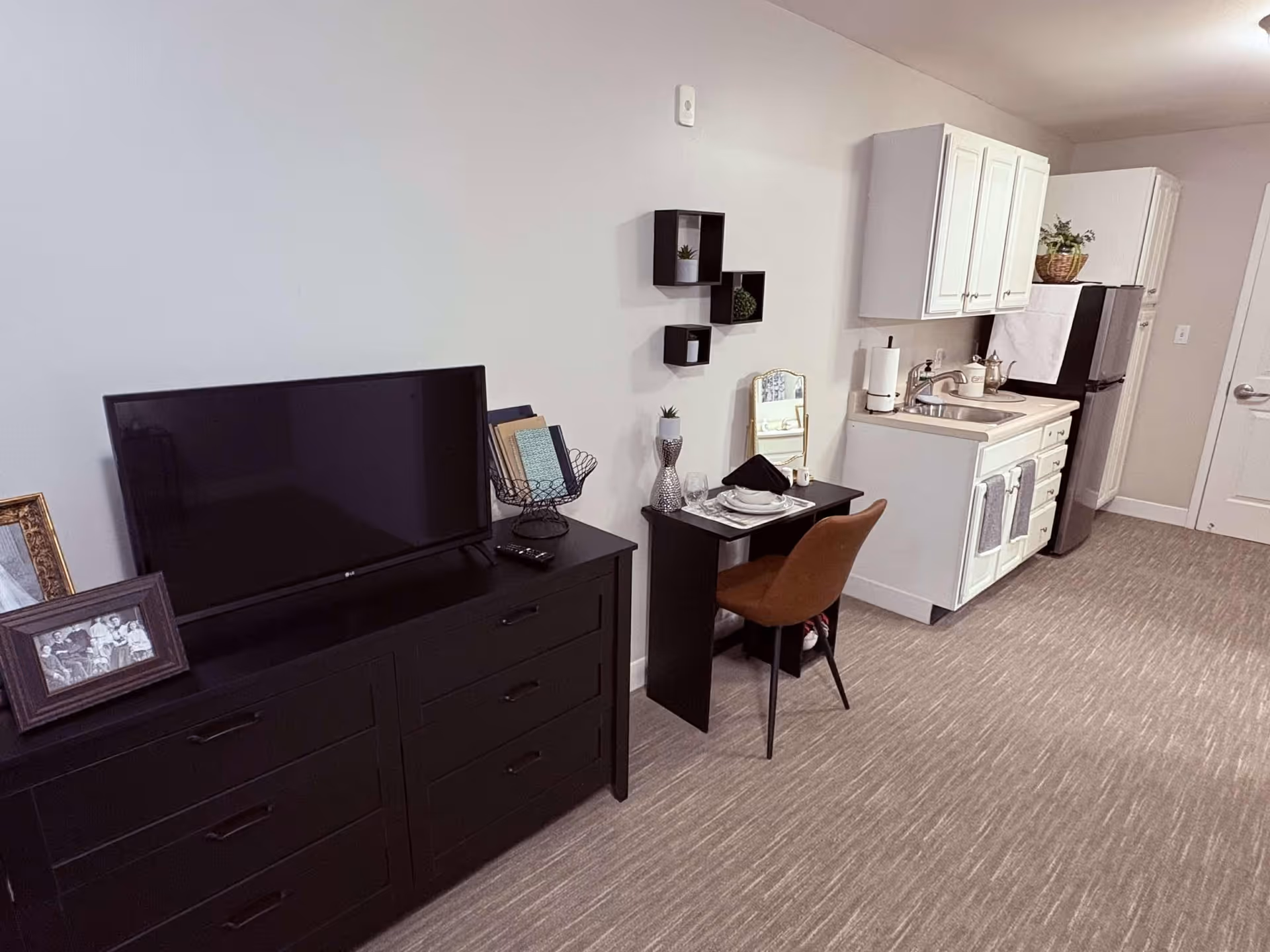 Interior view of a senior living facility room showing a black dresser with a flat-screen TV and framed photos on top, a small black desk set for one with a brown chair, and a kitchenette with white cabinets, a sink, and a refrigerator. The room has beige walls and carpeted flooring.