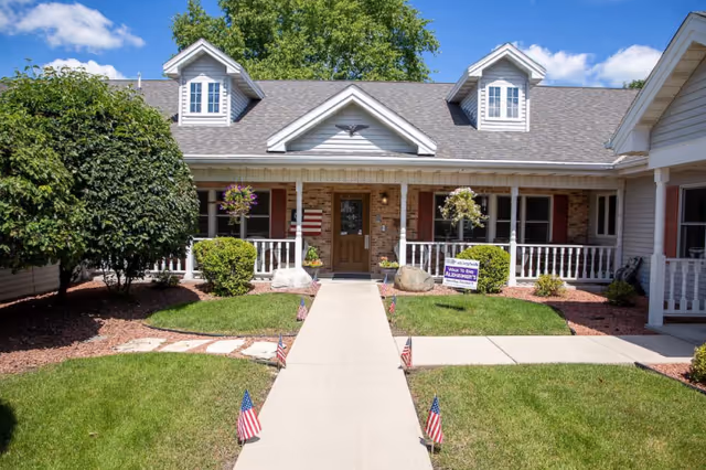 Front exterior view of a single-story assisted living facility building with a central entrance, white railings, hanging flower baskets, small American flags lining the walkway, and a sign indicating support for Alzheimer's awareness.