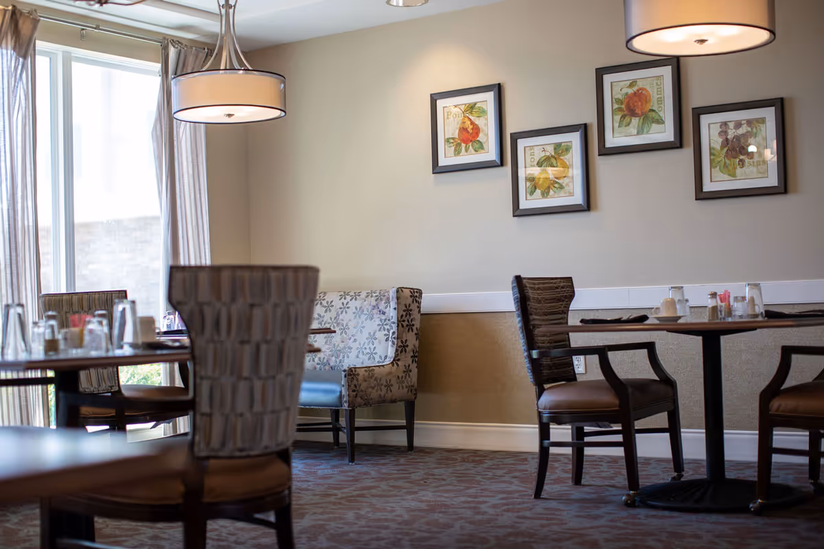 A dining area with several tables and chairs, including a cushioned bench with floral upholstery. The room has large windows with sheer curtains allowing natural light to enter. The walls are decorated with framed pictures of fruit. Overhead, there are modern pendant lights providing illumination.