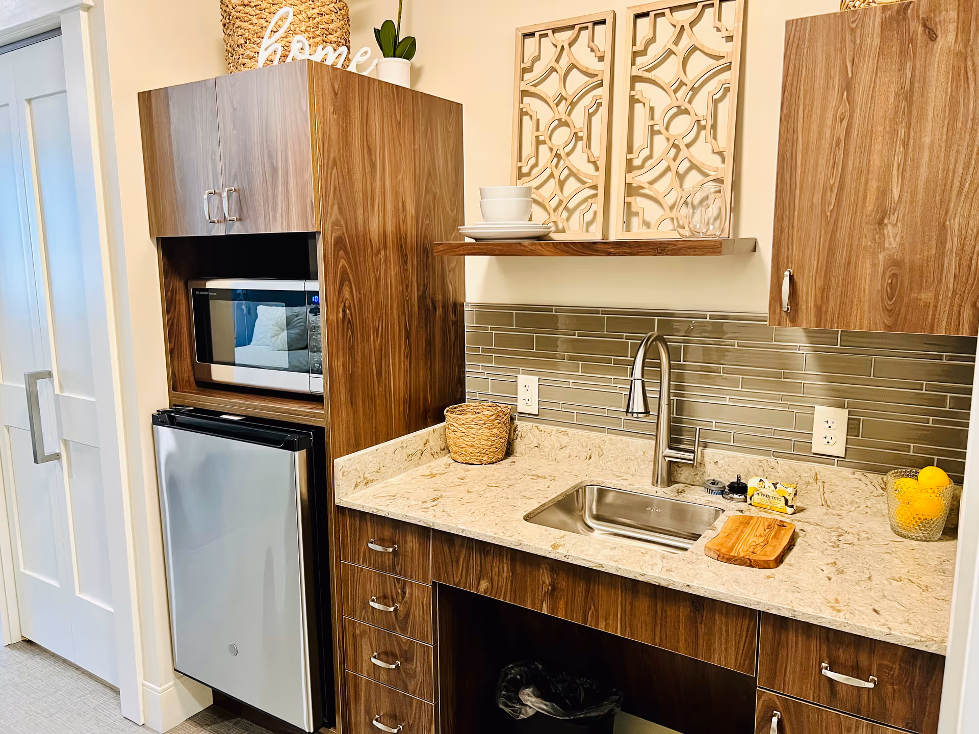 A compact kitchen area with wooden cabinets, a small stainless steel refrigerator, a microwave, and a sink with a modern faucet. The countertop is light-colored with a small cutting board, a basket, and a glass bowl filled with lemons. Above the sink is a shelf holding white dishes and decorative wooden wall art.