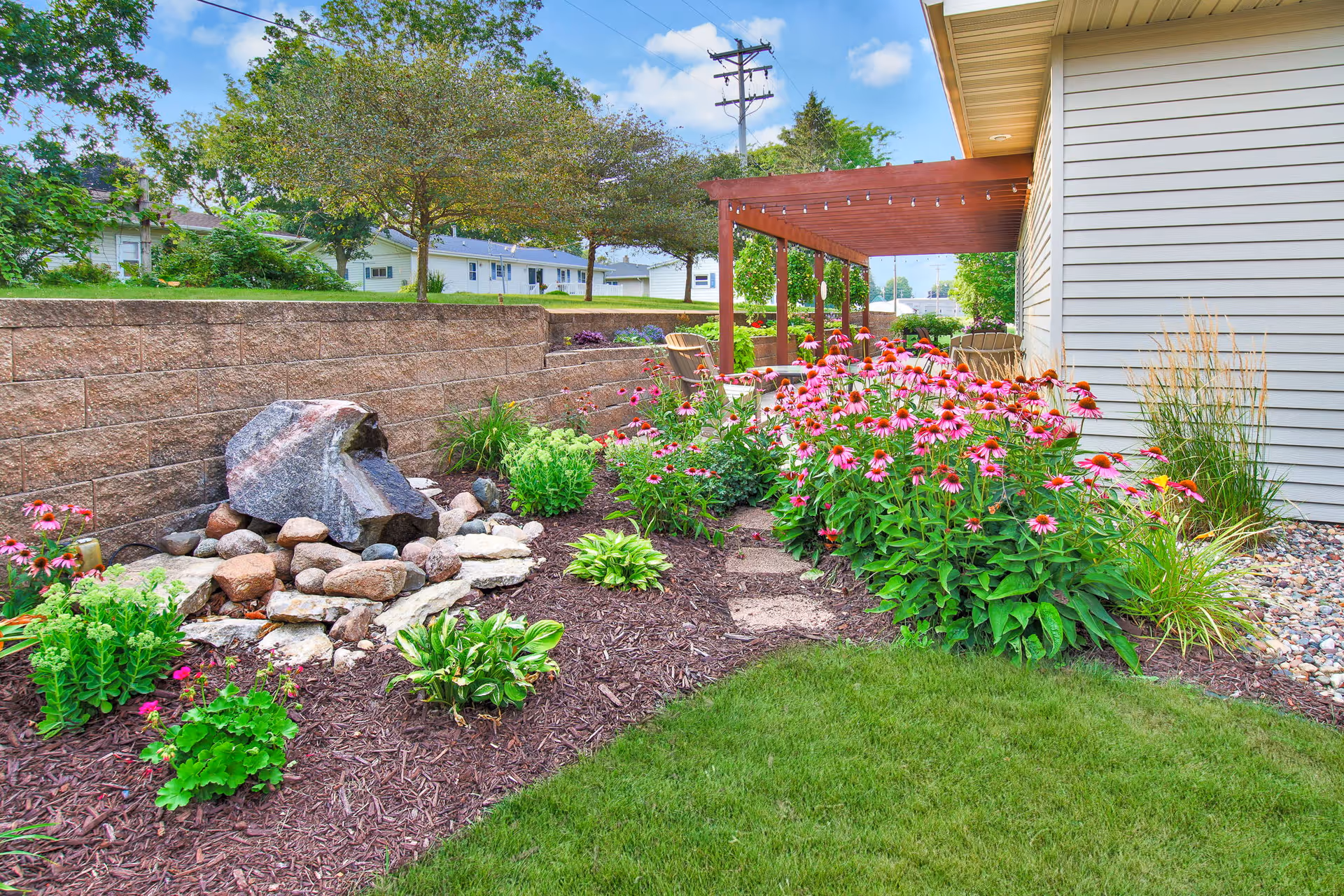 Landscaped outdoor garden with pink coneflowers, a rock water feature, pergola-covered patio and the side of a building.