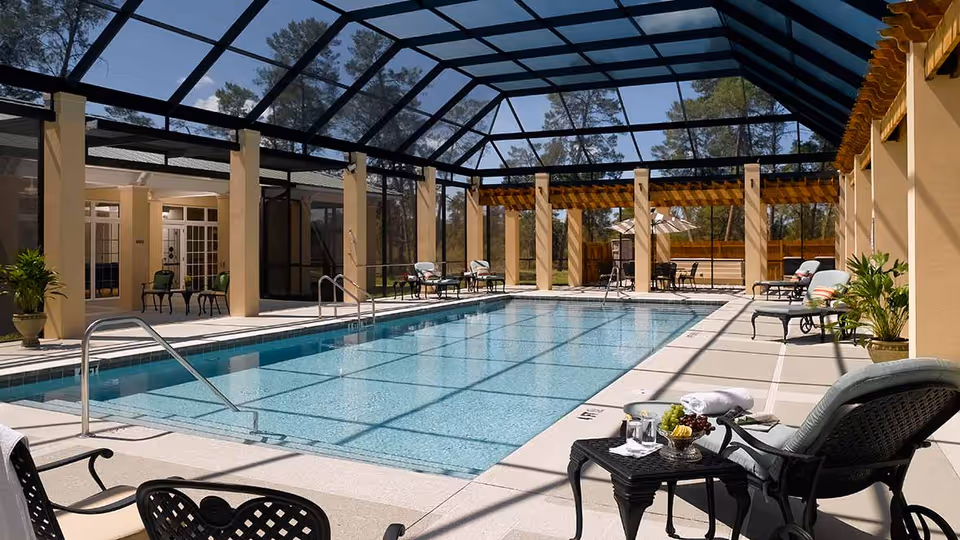 Indoor swimming pool area with a glass roof and beige pillars. Several lounge chairs and tables with chairs are arranged around the pool. There are potted plants and a small table with a towel, water glasses, and a bowl of fruit near one of the lounge chairs.