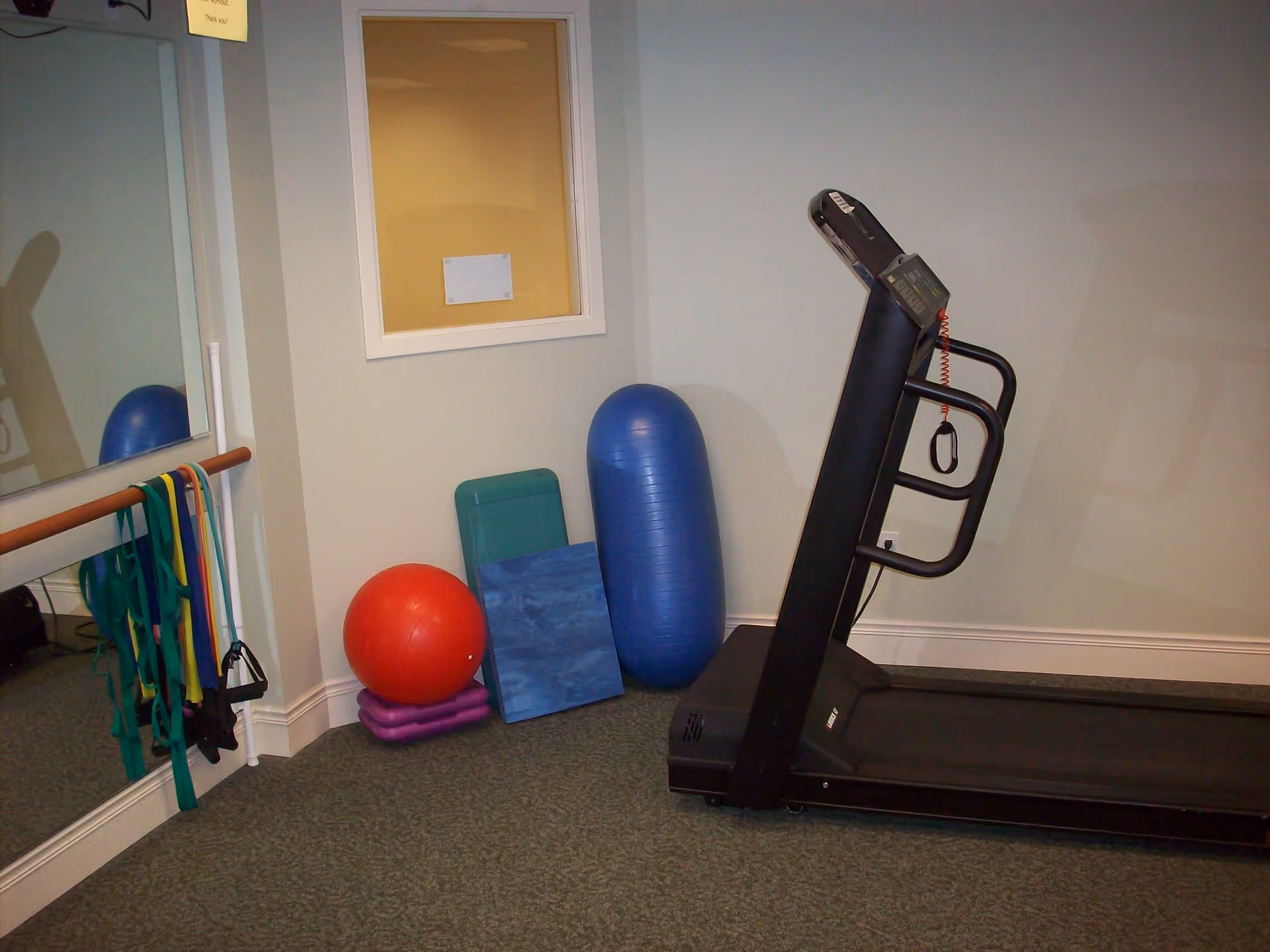 A small exercise room with a treadmill on the right side, various exercise equipment including resistance bands hanging on a wooden bar attached to a mirror, a red exercise ball, a blue balance cushion, and other fitness mats and blocks stacked against the wall.