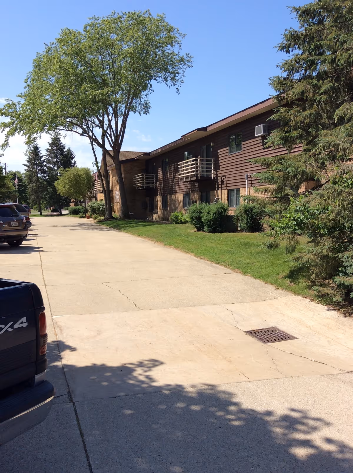 Exterior view of Simeon Square Retirement Community showing a two-story building with brown siding and small balconies. There is a paved driveway with parked vehicles on the left side and green grass with trees and shrubs on the right side under a clear blue sky.