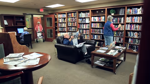 A senior living facility library with bookshelves filled with books along the walls. An elderly man is sitting in a black leather armchair reading a newspaper. An elderly woman is standing and looking at books on the shelves. Another elderly woman is walking with a walker, and a fourth person is seated at a computer desk. There are tables with newspapers and magazines in the room.