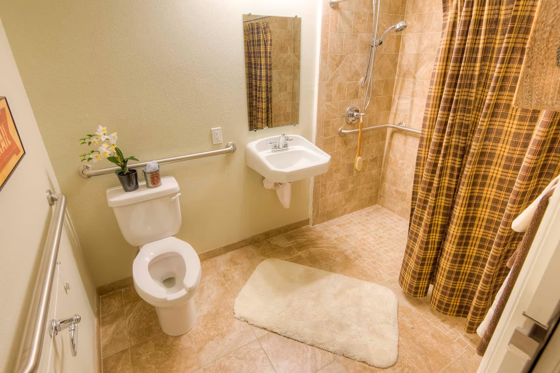 A bathroom with beige tiled floor and shower area. The shower has a plaid brown and yellow curtain and a handheld showerhead. There is a white toilet with a small potted plant and a candle on the tank. A white wall-mounted sink with a mirror above it is next to the shower. Grab bars are installed near the toilet and in the shower area. A white bath mat is placed on the floor in front of the sink.