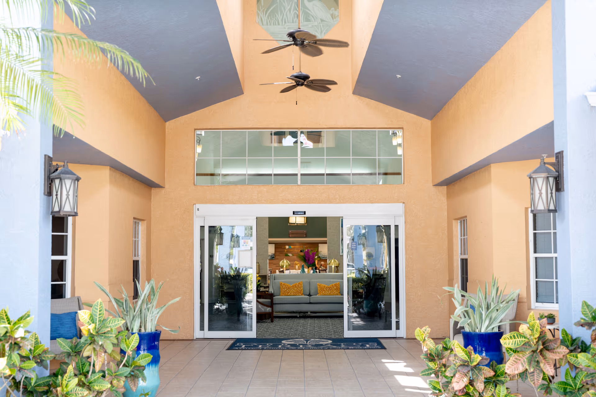 Entrance area of Barrington Terrace of Fort Myers featuring a covered walkway with ceiling fans, potted plants on either side, and glass sliding doors leading into a lobby with a sofa and decorative pillows.