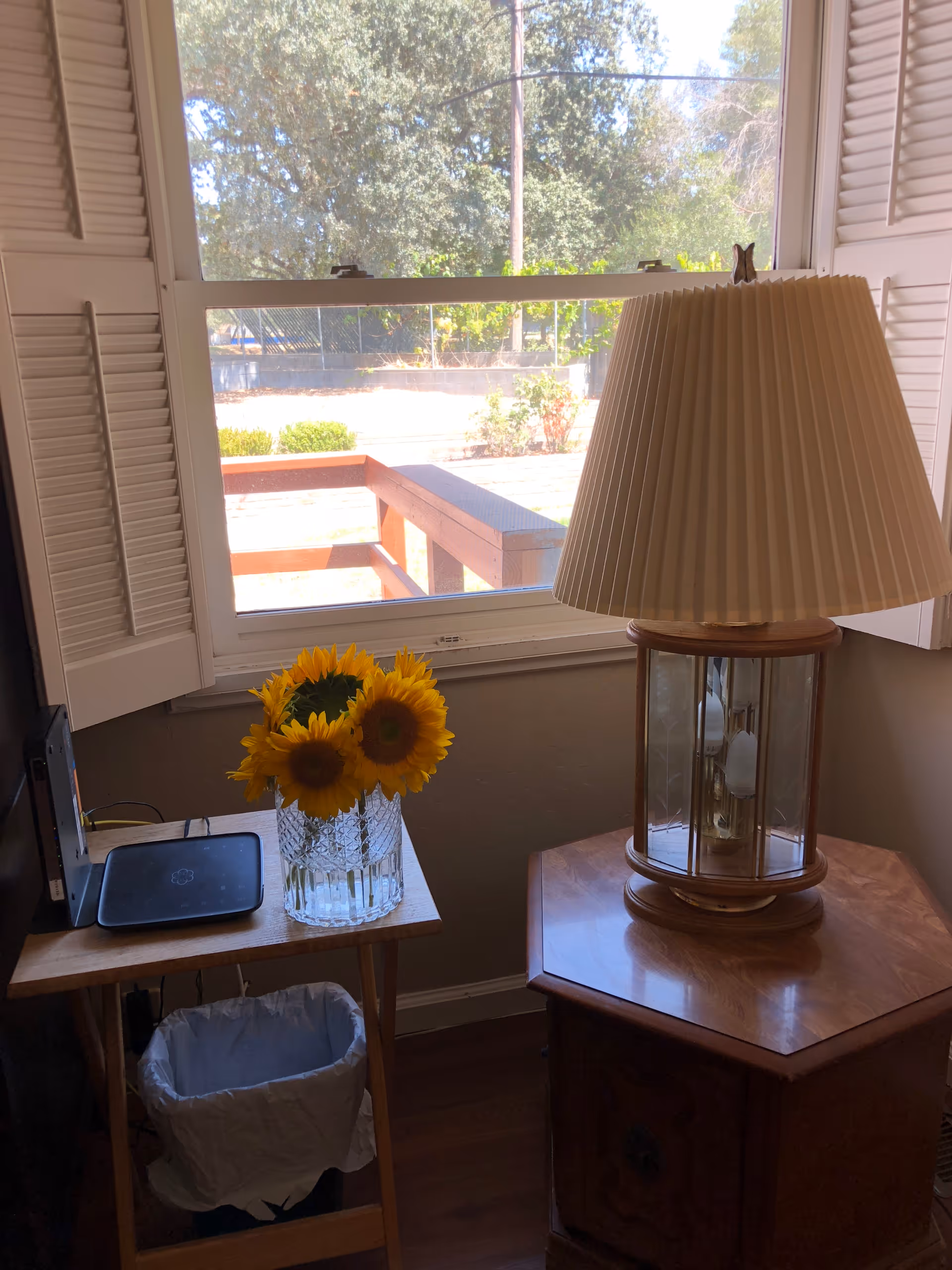 A cozy corner of a room with a window showing an outdoor view of trees and a wooden fence. Inside, there is a small wooden table with a glass vase holding bright yellow sunflowers, a black electronic device, and a trash bin underneath. Next to it is a wooden side table with a large lamp featuring a pleated beige lampshade.