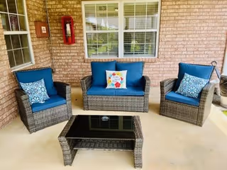 Outdoor patio area with a wicker furniture set consisting of two armchairs and a loveseat with blue cushions and decorative pillows, and a matching wicker coffee table with a glass top. The patio is adjacent to a brick wall with a window and a red fire extinguisher mounted on the wall.