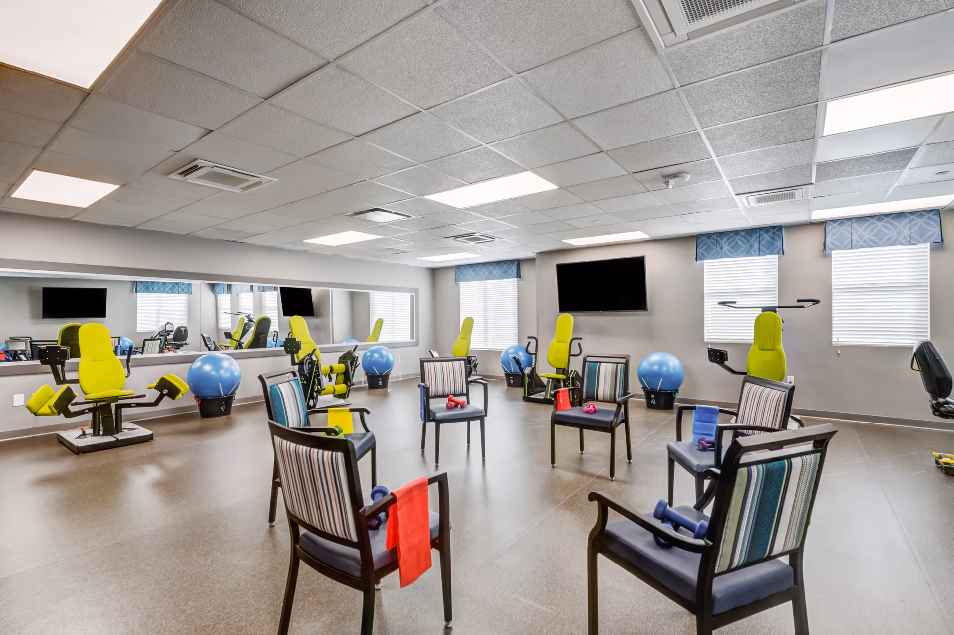 A bright exercise room with six chairs arranged in a circle, each chair holding small dumbbells and resistance bands. The room features several green exercise machines, blue exercise balls, a large wall mirror, two windows with blue valances, and a wall-mounted flat-screen TV.