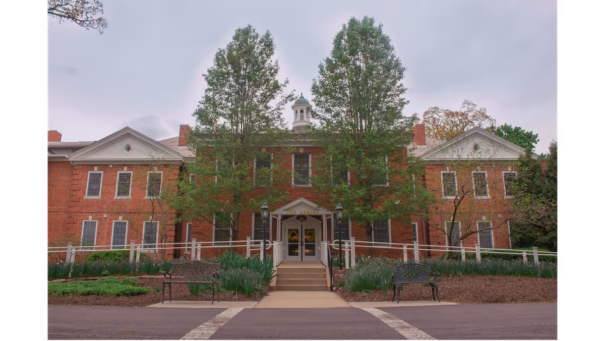 Front exterior view of a large red brick building with white trim, two tall trees flanking the entrance, a small set of stairs leading to double doors, and benches on either side of the walkway.