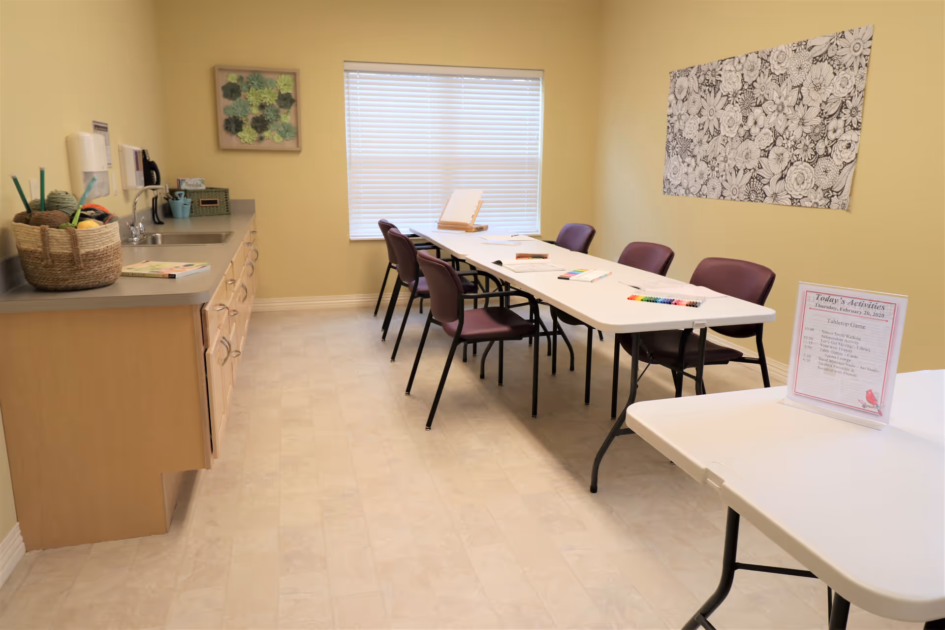 A bright activity room with a long white table surrounded by purple chairs. The table has coloring books, colored pencils, and markers on it. There is a countertop with a sink and a basket of yarn on the left side. The walls are painted light yellow and decorated with framed artwork. A sign on a smaller table displays today's activities.
