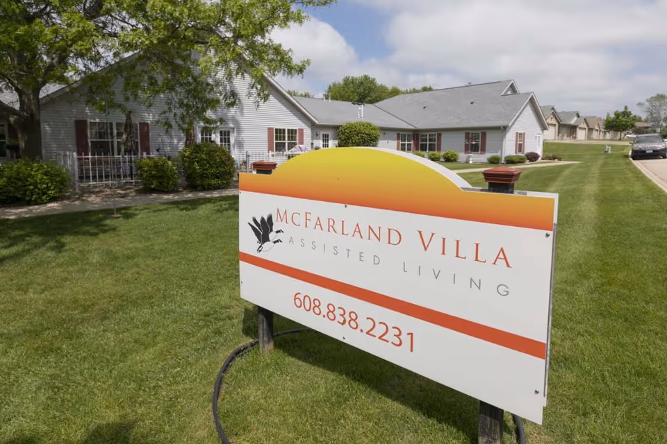 Outdoor view of McFarland Villa Assisted Living facility with a large sign in the foreground displaying the facility name and phone number. The building is a single-story structure with white siding and red shutters, surrounded by green grass and trees under a partly cloudy sky.